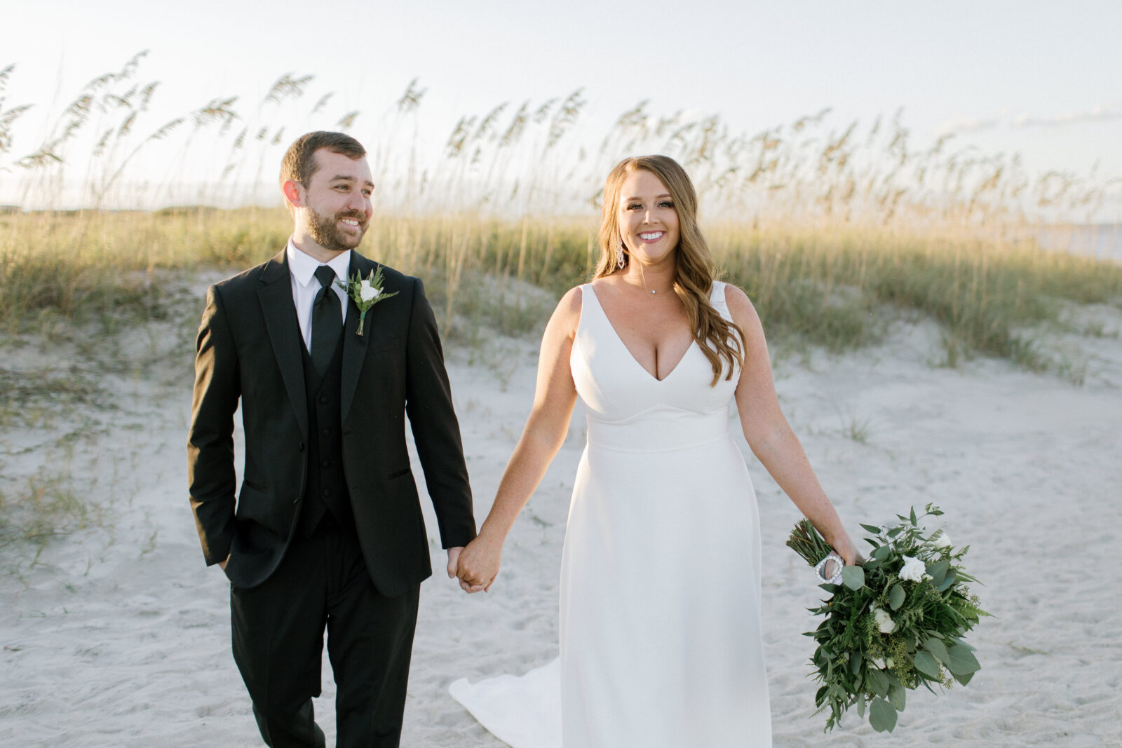 bride and groom walk on beach smiling with dunes in background