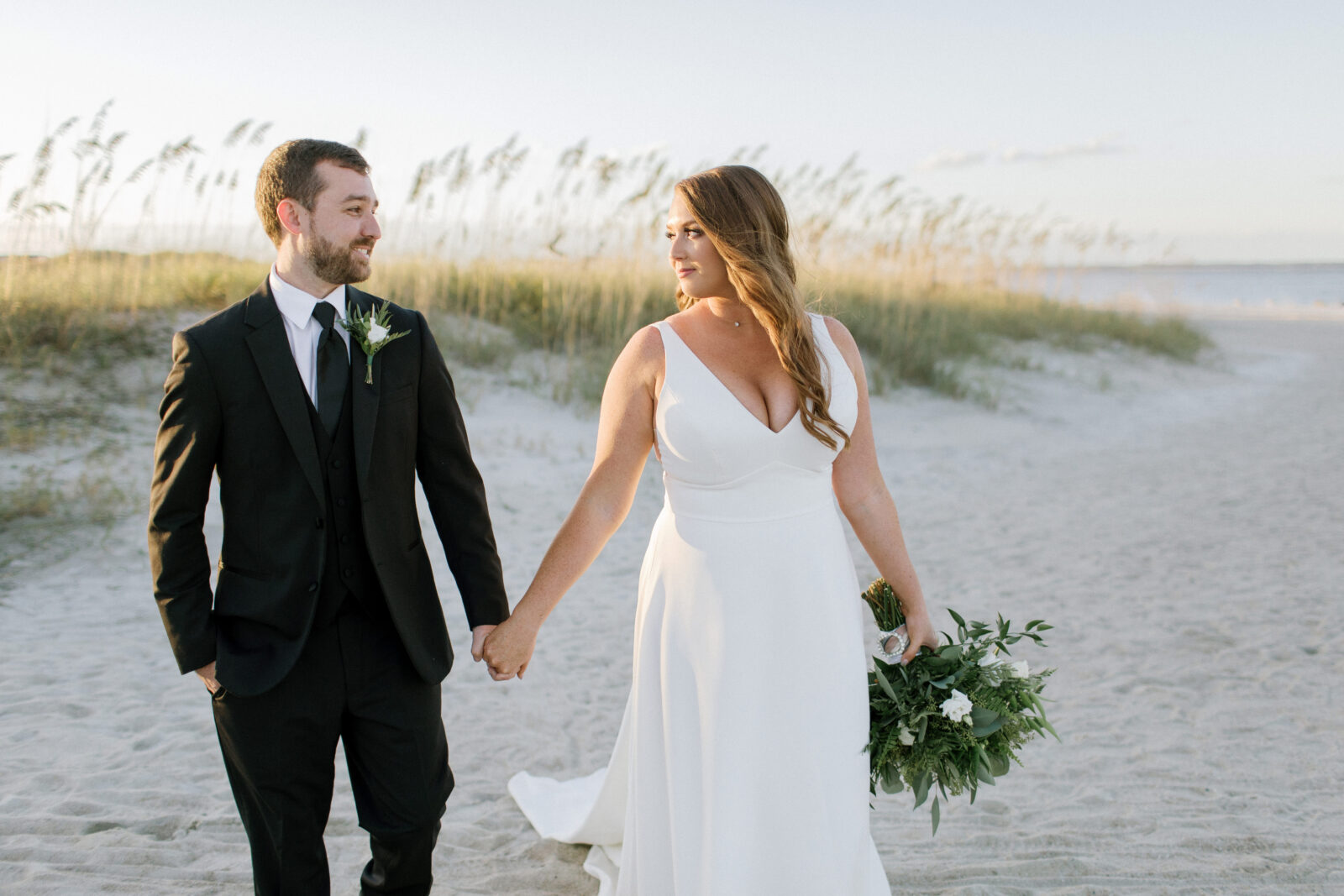 bride and groom walk on beach smiling at each other with dunes in background