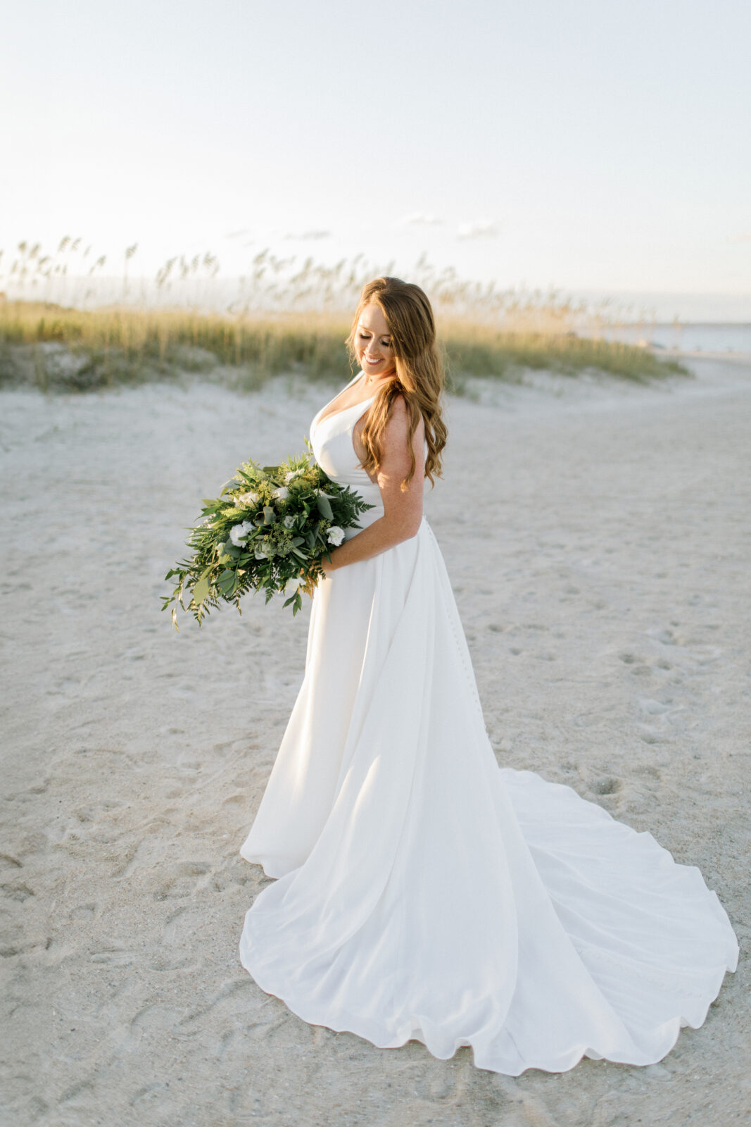 bride with long train holding bouquet and has red hair. dunes in background