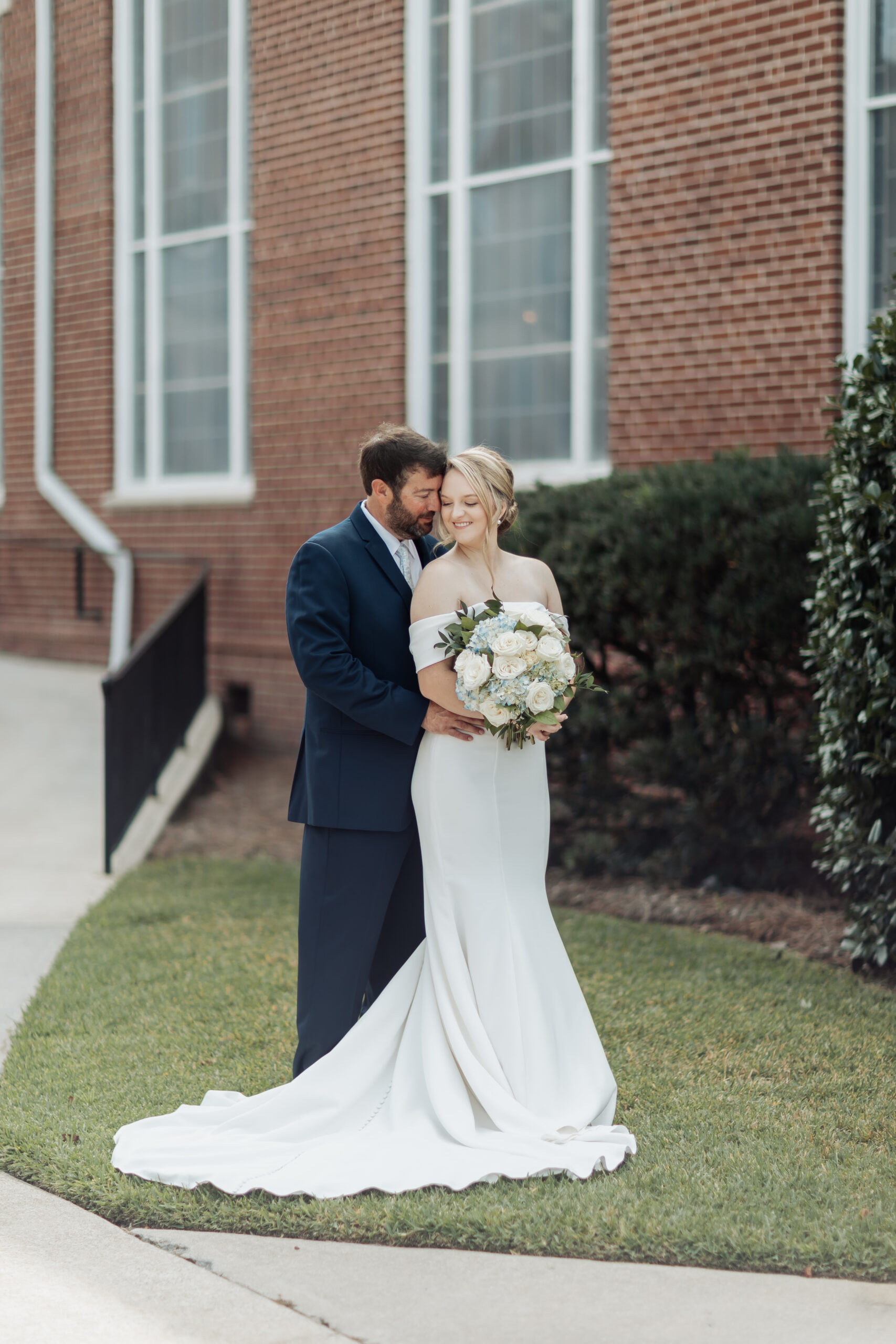 bride holding white flowers stands in front of groom in blue tux and looks back at him over her shoulder. They stand in front of brick church wall.